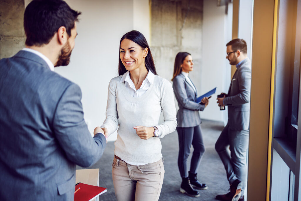 Friendly businesswoman shaking hands with her new colleague. In background are other two colleagues talking. Building in construction process interior.